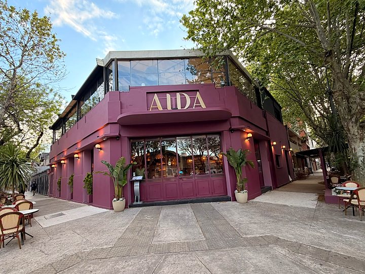 La fachada bordó de AIDA en la esquina, de día, con mesas en la vereda